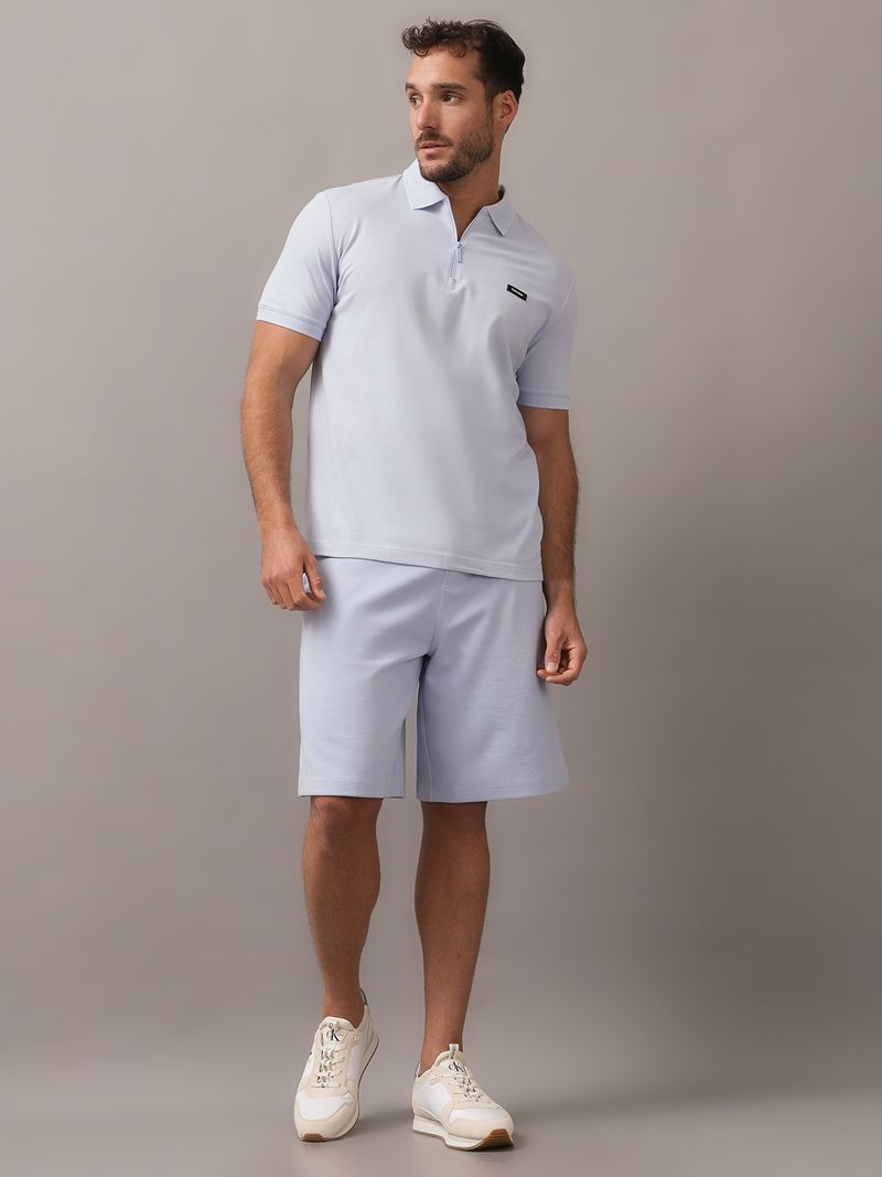 Hombre-con-polo-celeste-y-short-a-juego-de-Calvin-Klein-posa-con-una-mano-en-el-bolsillo-y-otra-en-el-cuello