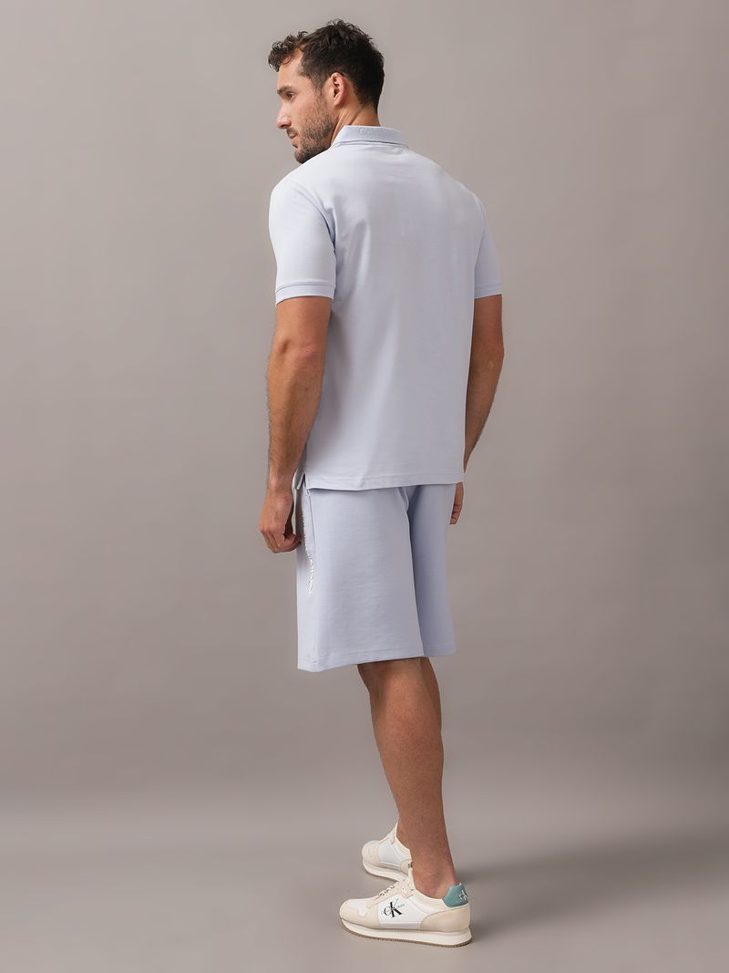 Hombre-con-polo-celeste-y-short-a-juego-de-Calvin-Klein-posa-con-una-mano-en-el-bolsillo-y-otra-en-el-cuello